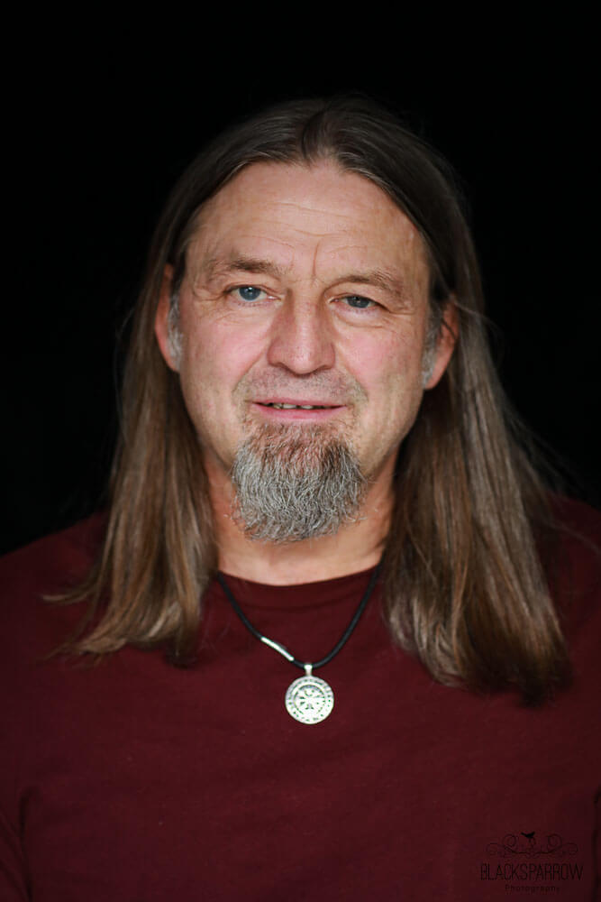 Porträt eines Mannes mit langen Haaren und grauem Bart, der ruhig in die Kamera blickt.