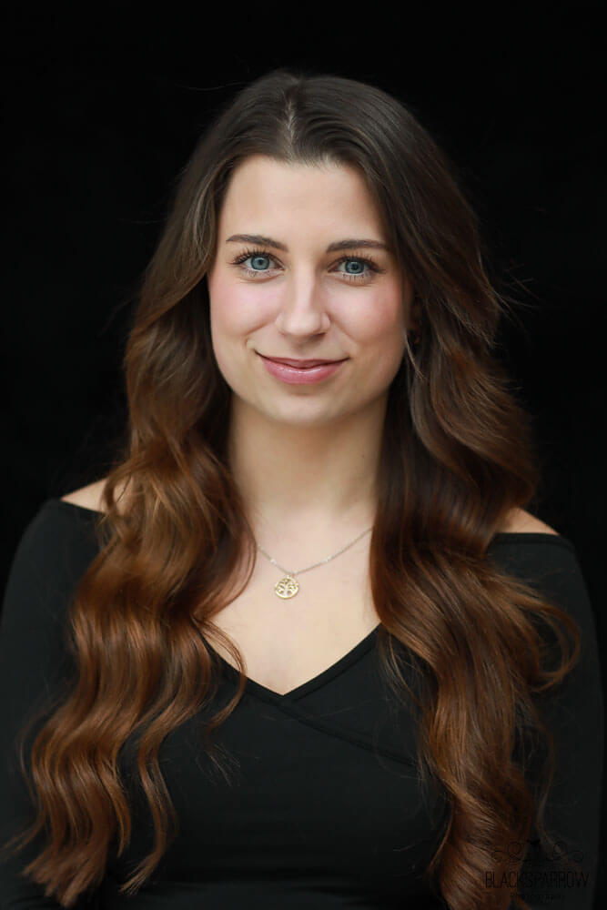 Porträt einer jungen Frau mit langen braunen Haaren, die lächelnd in die Kamera blickt, neutraler Hintergrund.